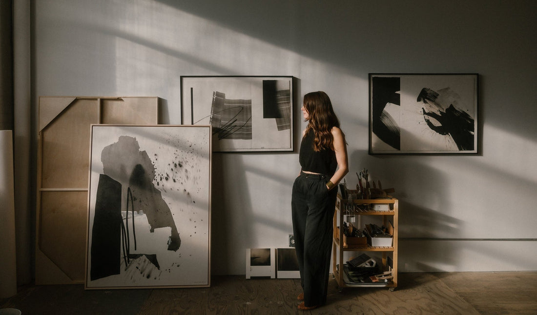 Artist standing in her studio looking out window with 3 pieces of her abstract art in the background
